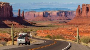 A white SUV with gear on its roof travels down a winding road through the Desert Southwest, passing red rock formations, mesas, and tall cacti. The sky is partly cloudy and distant mountains rise in the background, evoking a classic road trip adventure.